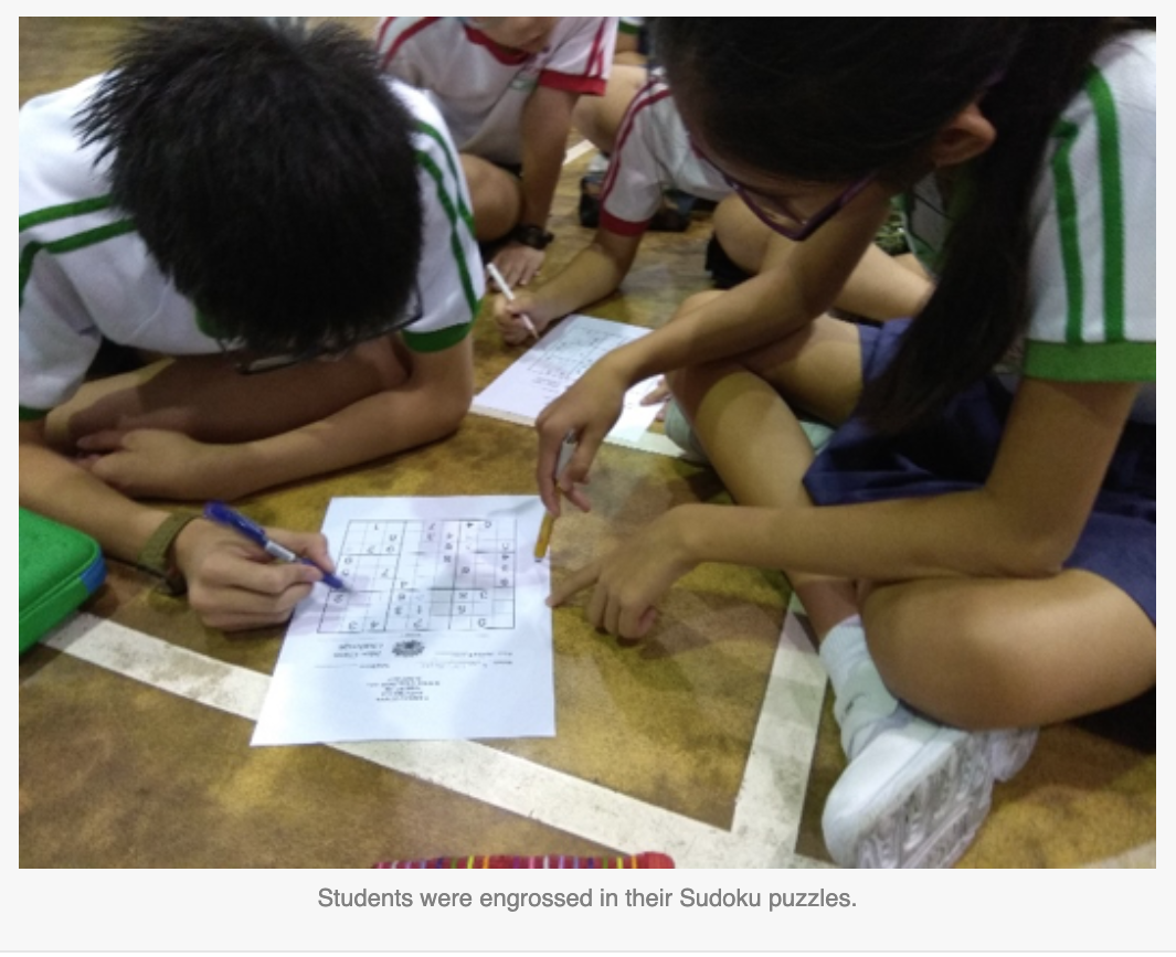 Several students in white and green uniforms sit on a floor, writing on printed Sudoku puzzle sheets.